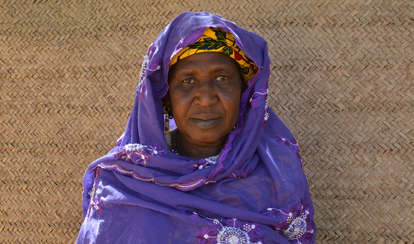 A close up of Aminatou wearing a purple shawl standing in front of a beige-coloured wall.