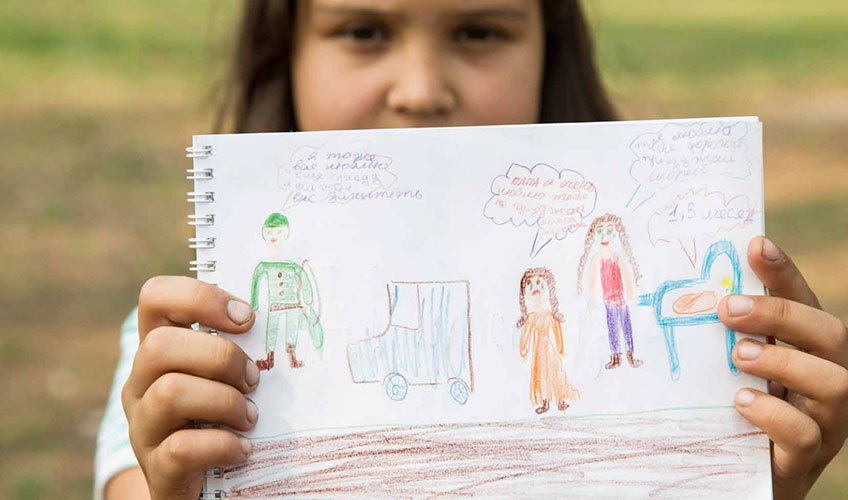 A young girl at a Peace Education camp holds up a drawing of people with speech bubbles and Russian text