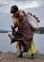 A woman at a riverbank with a bundle of chickens on her back