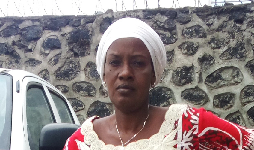 Odette Budari Kamanzi leans on a white car wearing a red and shite outfit, standing in front of a stone wall.