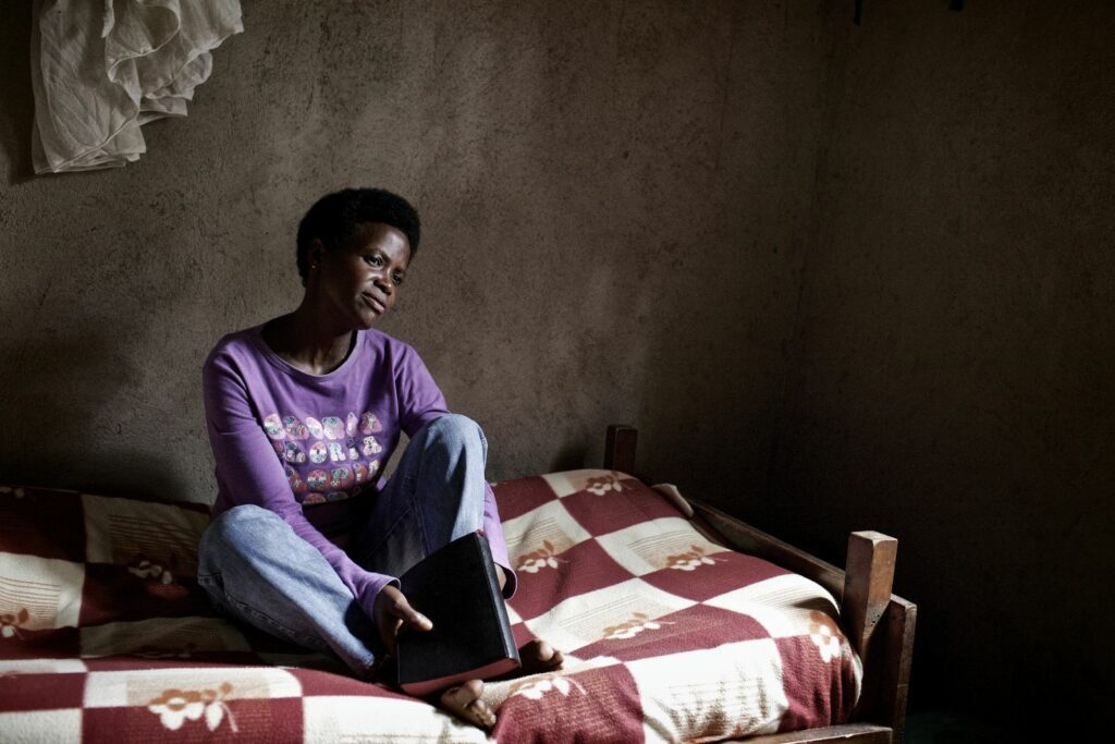 Claudine sits on her bed wearing a purple long-sleeved top and blue jeans. The bed has a red and white checked cover. 