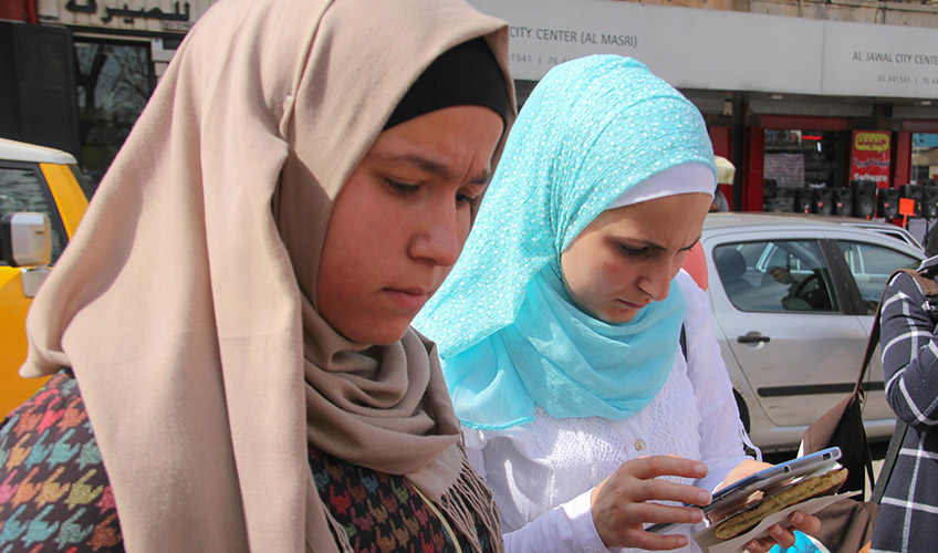 Two women map points on their phone. A woman on the left wears a beige hijab and the woman on the right wears a light blue hijab and looks at her phone.