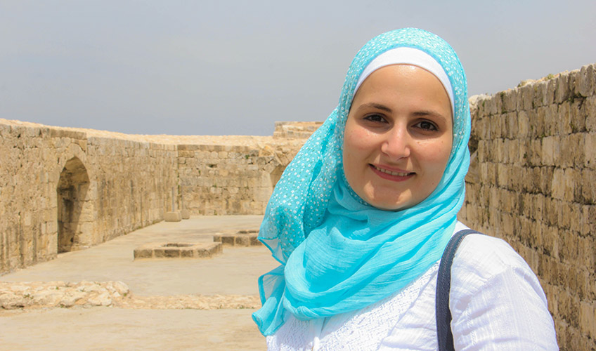 Ifdal wears a light blue hijab and white top. She stands in front of a yellow wall leading into the distance. 