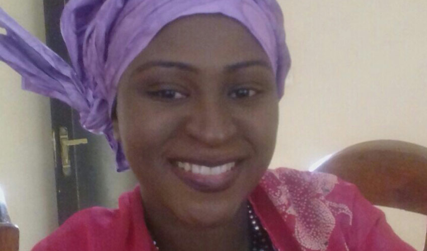 A close up of a woman smiles. She is wearing a purple headscarf and red top. 