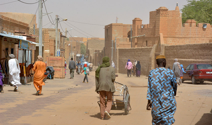 A dusty orange street scene in Mali with people walking. 