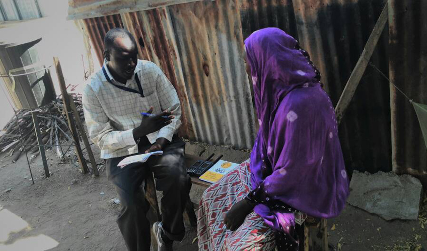 A man in a shirt holding a phone speaks to a woman in a purple shawl.