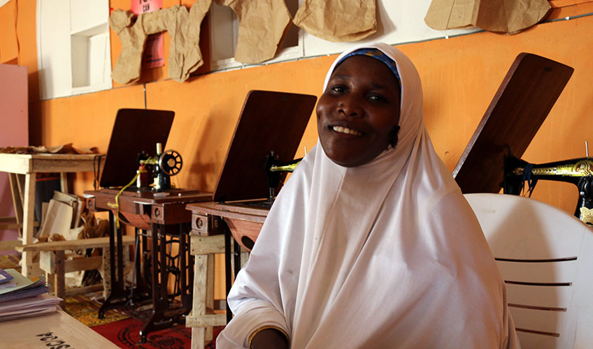 Fatima smiles wearing a white hijab in front of an orange wall surrounded by sewing machines.