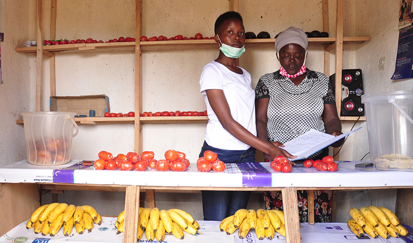 Julliene and her daughter stand to the right of their , Mauelle, at the market selling their goods at the market. They stand behind a market stall covered with tomatoes and bananas.