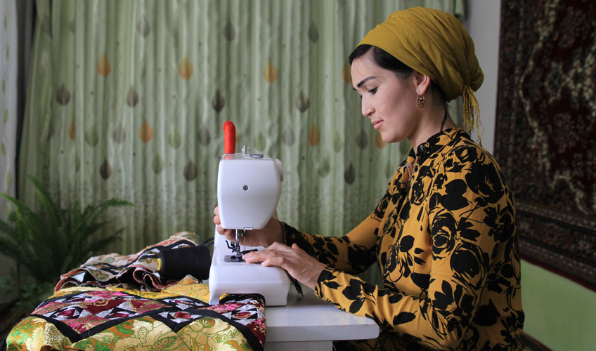 A woman in a yellow and black dress and yellow headscarf works at a sewing machine. A green patterned curtain hangs in the background.