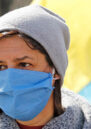 A woman wearing a blue masks and grey woolly hat with a flag of Ukraine in the background.