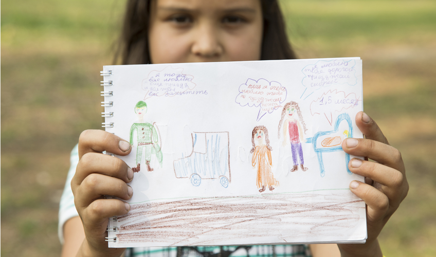 Girl holds up a drawing of epople with speech bubbles and Russian text