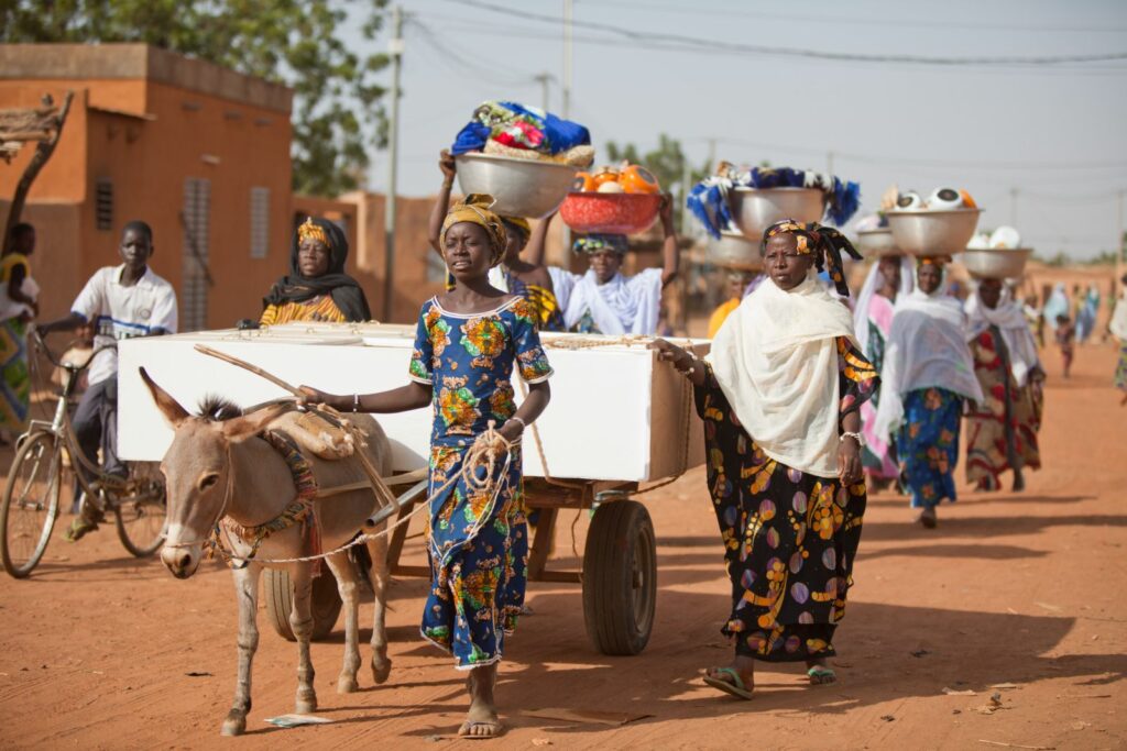 A procession of wedding guests walk along with a donkey and cart on an earthen red road with gifts and baskets.