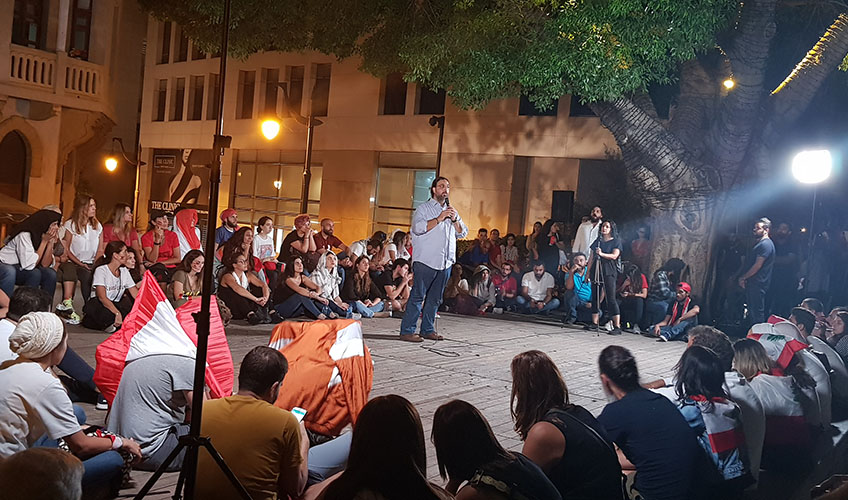 A group of people sitting on the ground in a square while a man with a microphone stands in the middle. ut, October 2019