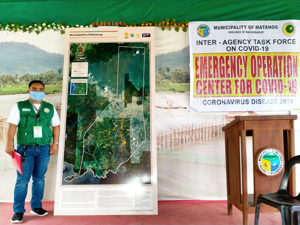 A man in a mask and green hazard jacket stand in front of signage from the mapping project.