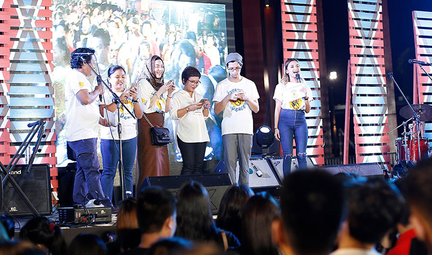 A group of young people on stage looking at an app on their phones and smiling as the announcer speaks