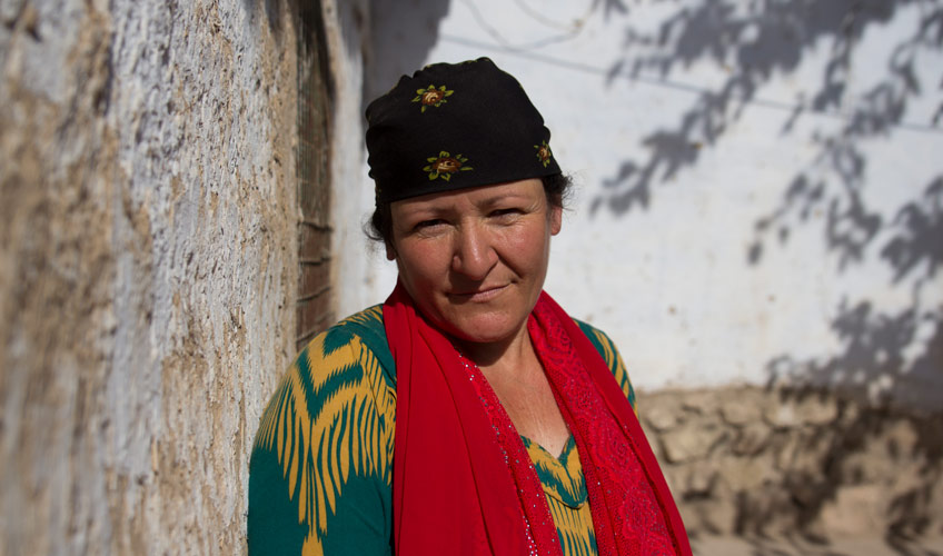 A woman with a neutral expression in a green and yellow jumper, red scarf and black hat stands beside a tree. 