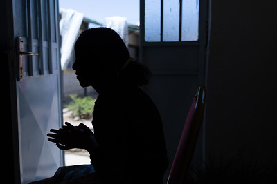 Photograph of young woman, seen as a silhouette, sitting in on a chair front of a door. Through the door you can see an IDP (Internally Displaced People) camp. 