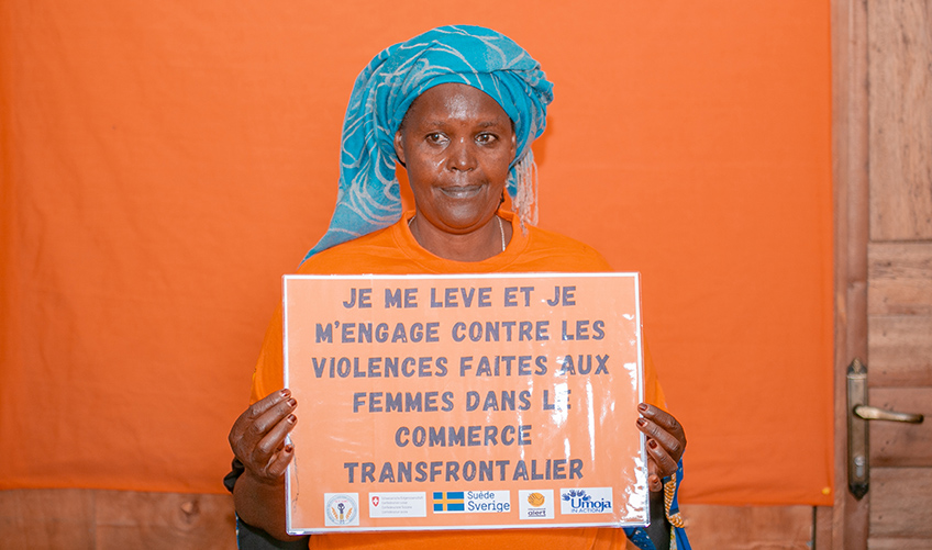 A woman wearing orange poses against an orange wall with a sign in French vowing to combat violence against cross border traders