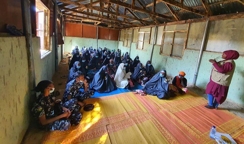 A group of women sat on the floor, most wearing traditional hijab and two in the foreground in uniform.  A woman is speaking to the gorup.