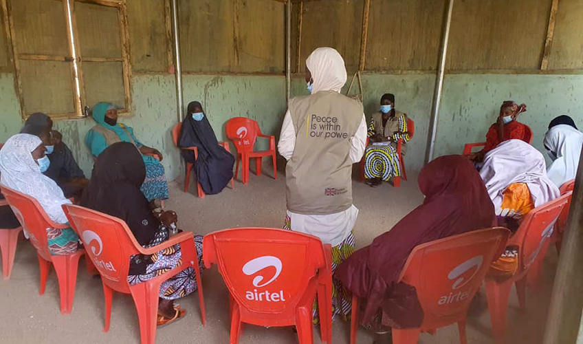 Women in hijab and wearing face masks seated on chairs in a circle listening to a woman standing in the centre.