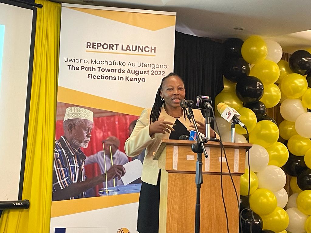 Image shows Leah standing at a podium at the Uwiano, machafuko au utengano report launch