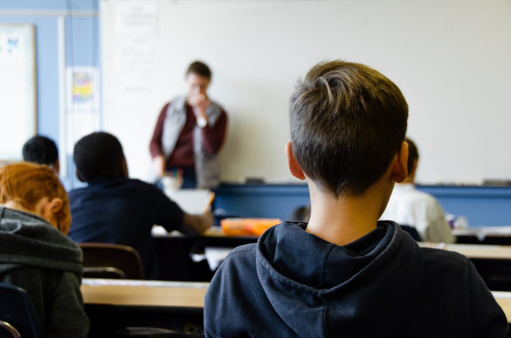 Image shows students sitting in a classroom looking at a teacher, who is standing next to a whiteboard.