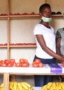 Julliene and her daughter stand to the right of their , Mauelle, at the market selling their goods at the market. They stand behind a market stall covered with tomatoes and bananas.