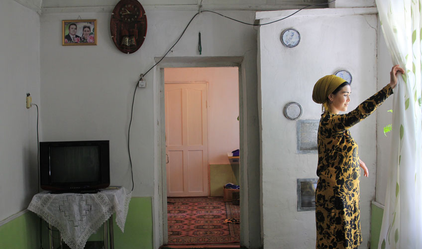 A woman in a black and yellow dress draws back a curtain at the window.