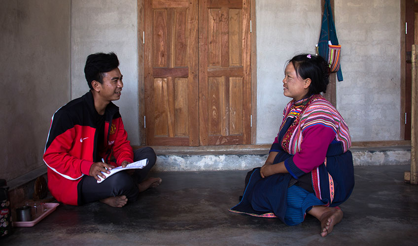 A man and woman sit on the floor in front of a brown door. The man is smiling and wearing a red and black jacket, the woman is wearing a pink jacket and blue trousers.