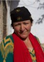 A woman with a neutral expression in a green and yellow jumper, red scarf and black hat stands beside a tree.